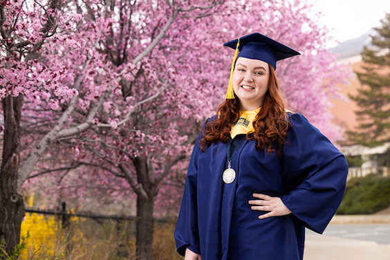 Student graduation speaker Emilee Carr poses in cap and gown by a light gray backdrop.