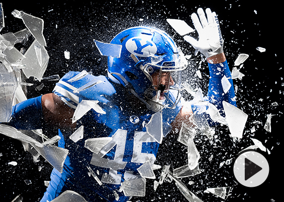 A BYU football player in a royal blue uniform and helmet runs through a sheet of sugar glass.