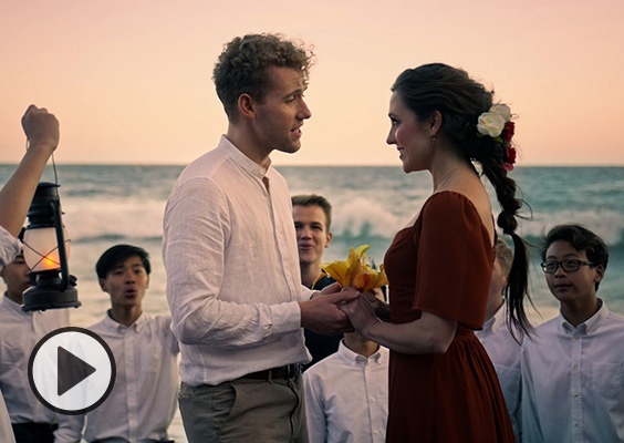 A BYU Vocal Point singer with curly blonde hair and Cinderella aka Laura Osnes lock gazes and sing a duet at the seashore as the All-American Boys Chorus hold lanterns and join in the song.