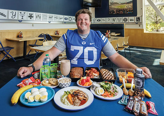 A football player is surrounded by the food he eats in a day, including eggs, oatmeal, smoothie, meat, fruit, and protein bars.