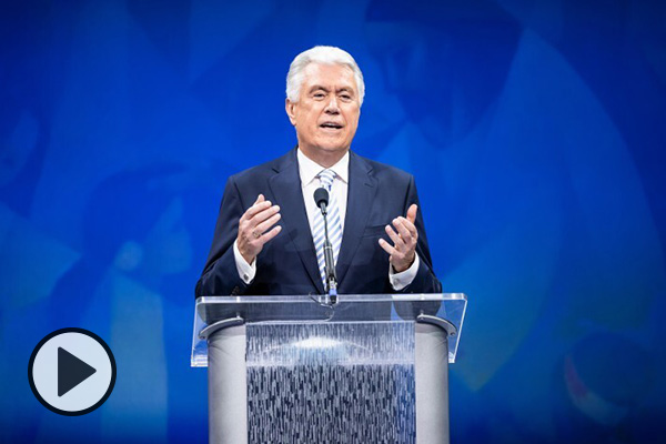 As part of Education Week, Elder Dieter F. Uchtdorf spoke at a devotional. Here he stands at a podium in the Marriott Center framed by a blue background.