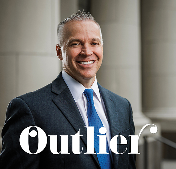 BYU President Shane Reese in a dark suit and blue tie smiles near the columns of the Maeser Building. The text says Outlier.