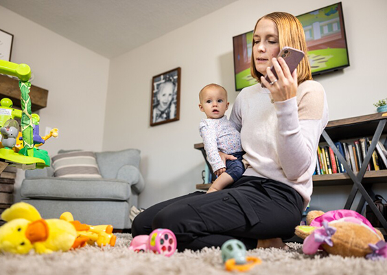 A mom holds her phone and a baby in a room filled with books and toys to illustrate a study that finds that moms shoulder disproportionate demands from kids’ schools. Photo by Rebeca Fuentes/BYU Photo.