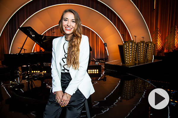 Singer Lauren Daigle smiles in front of a stage with a curtain, music stands, and circular amber lighting.