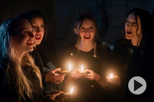 BYU Noteworthy members sing while holding candles and wearing cloaks against a Bible set.