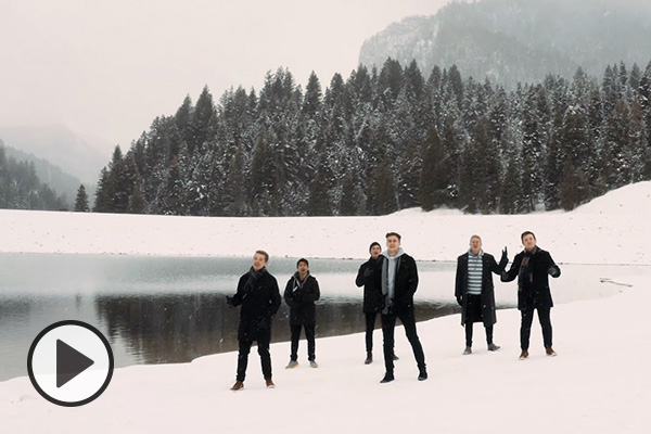 BYU Vocal Point members in dark winter clothing sing from a snowy pine forest near a lake.