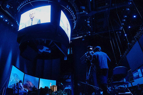 A speaker, multiple screens, and the LED board, along with all of the elements of a BYU devotional being held in Marriott Center. A TV camera is shown in the foreground.