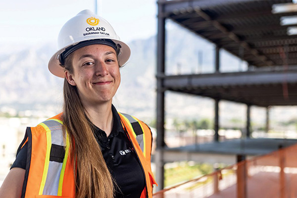 BYU student Savannah Taylor is wearing a construction hat and orange vest. She smiles as she stands near steel supports and scaffolding. Photo by Brad Slade.
