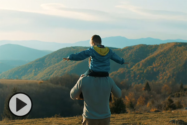 In a meadow above the mountains, with sky and clouds visible for miles, a young boy extends his arms as he is given a ride on his father's shoulders.