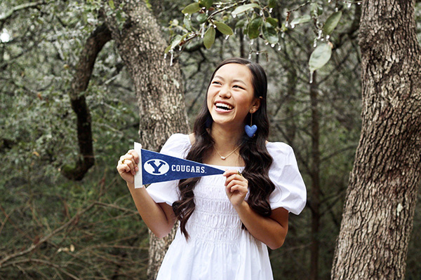 New BYU student Hannah Larson is wearing a large blue heart earring and a huge smile as she stands in a wooded area, holding a blue and white Y Cougars banner. Photo by Kelly J. Larson.