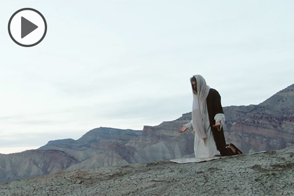 An actor portraying Jesus Christ kneels on a slope overlooking a mountainous desert.