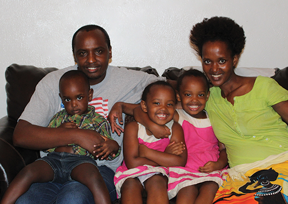 A refugee family with three young children smiles and cuddles on the couch.