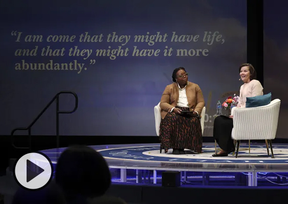 Primary General President Susan H. Porter and Sister Tracy Y. Browning on stage at BYU Women’s Conference. A screen displays a scripture quoting Jeus Christ. “I am come that they might have life, and that they might have it more abundantly.”