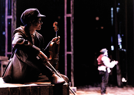 On a stage during a live performance of the Fiddler on the Roof, a female student sits in the foreground holding a violin, gazing at an actor in the background speaking to an audience.