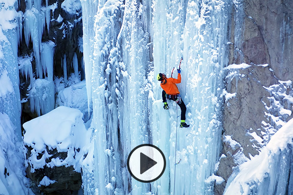 A climber in an orange jacket uses ice climbing gear to ascend a frozen waterfall.