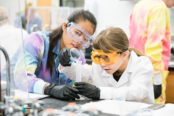 A young woman in white lab clothes, goggles, and gloves pours liquid from a glass container into a beaker while an older female labworker assists.