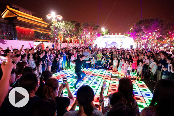 A hoop dancer performs in a public space in China at night. He is surrounded by a crowd taking pictures and cheering.