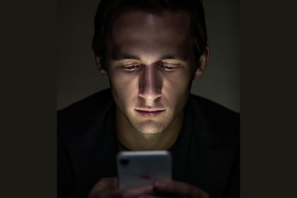 A smartphone user looks intently at his device whick illuminates his face against a dark background.