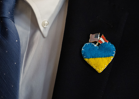 A closeup of a blue tie, white shirt and dark suitcoat. Pinned to the lapel is a blue and yellow heart, the colors of the Ukrainian flag.