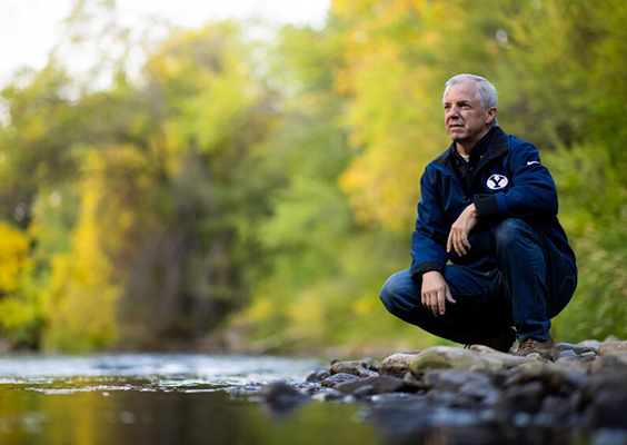 BYU civil engineering professor Jim Nelson, a hydrologist, kneels next to a river bank, green forest foliage in the background.