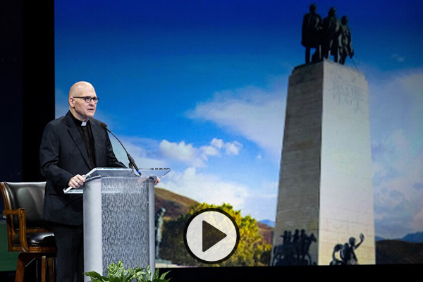 Rev. Dr. Andrew Teal stands at the Marriott Center podium, flanked by a projected image of This Is the Place Monument.