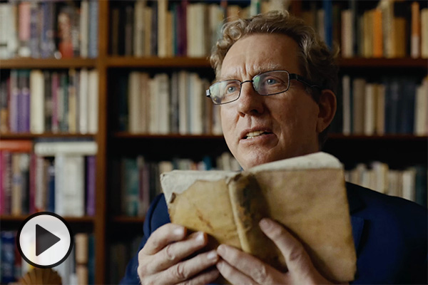 Spanish lit professor John Rosenberg holds up a book in front of multiple library shelves.