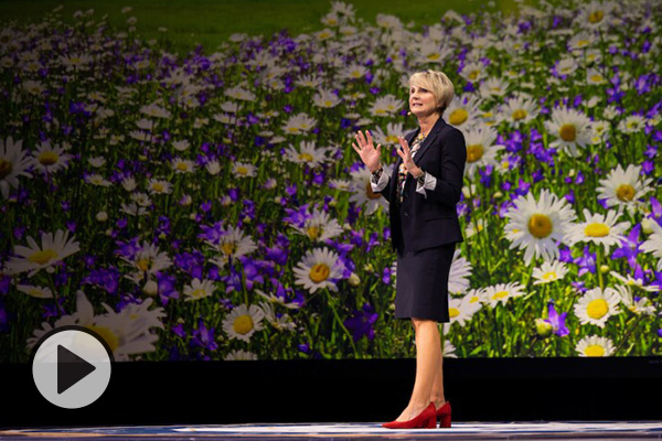 Julie Valentine gives a devotional address, a field of flowers displayed on a screen in the background.