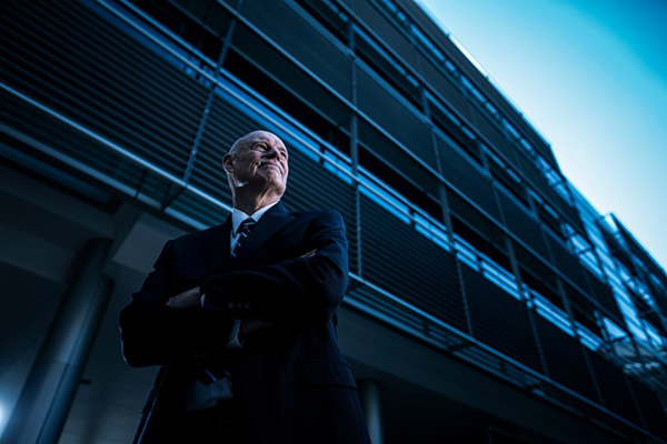 Forensic linguist William Eggington, arms folded and wearing a dark suit, stands in front of a tall building wrapped in metal railings.