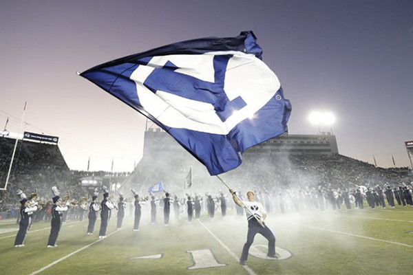 In a cloud of white smoke, a male BYU cheerleader waves a Y Flag on the football field in front of a line of marching band members.