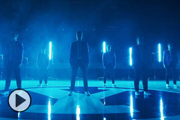 Bathed in blue light and wearing blue suits and ties, BYU Vocal Point members stands socially distanced on a high-gloss wooden playing floor, a Y logo at center.