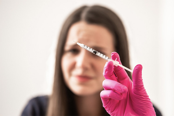 This photo focuses on a pink-gloved hand holding a loaded syringe. A female with long brown hair looks concerned in the background nearby.