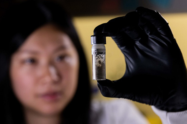 A black-gloved student researcher in a white lab coat holds up a test sample.