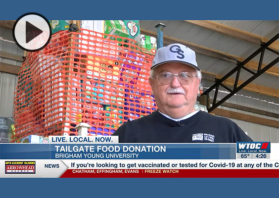 A gentleman from Georgia stands in front of a wrapped pallet of donations in a warehouse as he is interviewed about a BYU tailgate food donation.