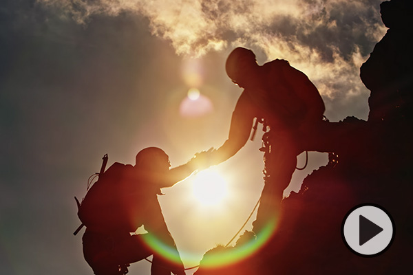 A climber extends a hand to another below him on a craggy, sunlit cliff.