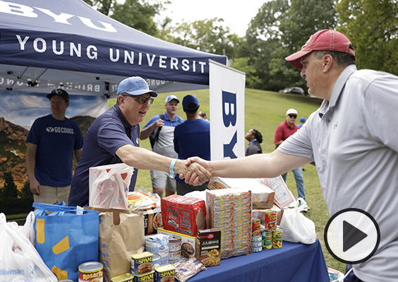 A BYU tailgater shakes hands with an Arkansas fan over a table stacked with food donations.